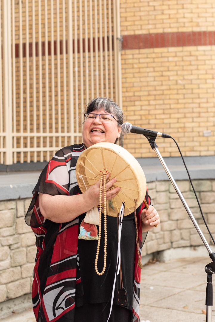 Hand Drum Singing Time with Brenda MacIntyre Brenda MacIntyre, Medicine Song Woman, laughing and holding her hand drum outside with a microphone.