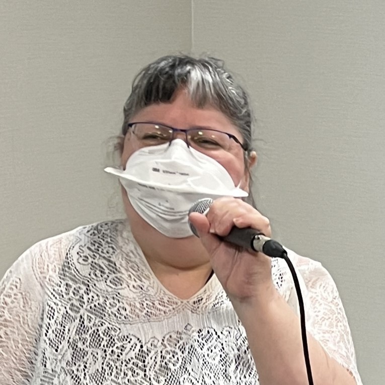 Brenda MacIntyre at the mic wearing a white shawl and Vflex N95 mask